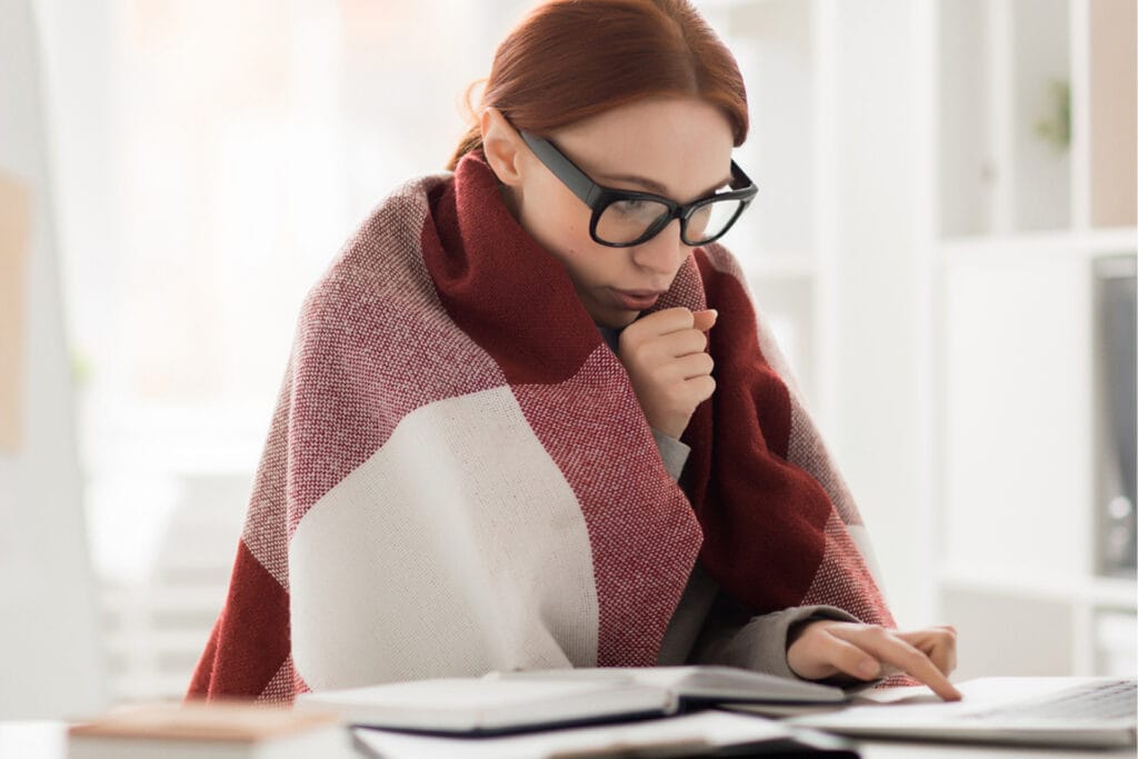 Woman wrapped in blanket while blowing warm air into her hand at her table inside.